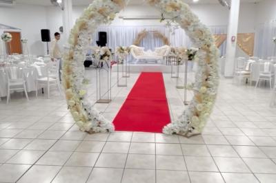Salle de réception décorée pour un mariage, avec tables dressées, fleurs blanches et rouges, et arche florale en arrière…