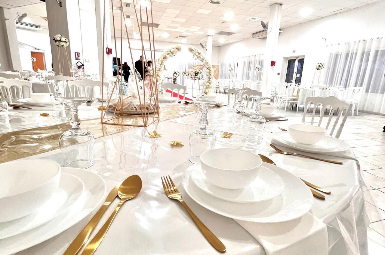 Table de réception élégamment dressée avec vaisselle blanche, couverts dorés et décoration florale dans une salle lumineuse.