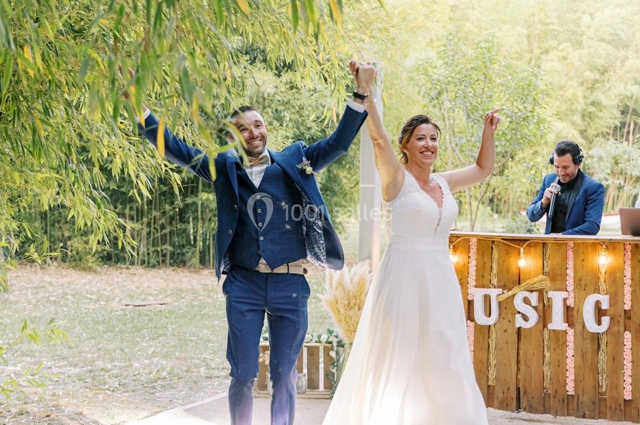 Un couple en tenue de mariage lève les bras en souriant, devant un décor champêtre avec un DJ en arrière-plan.
