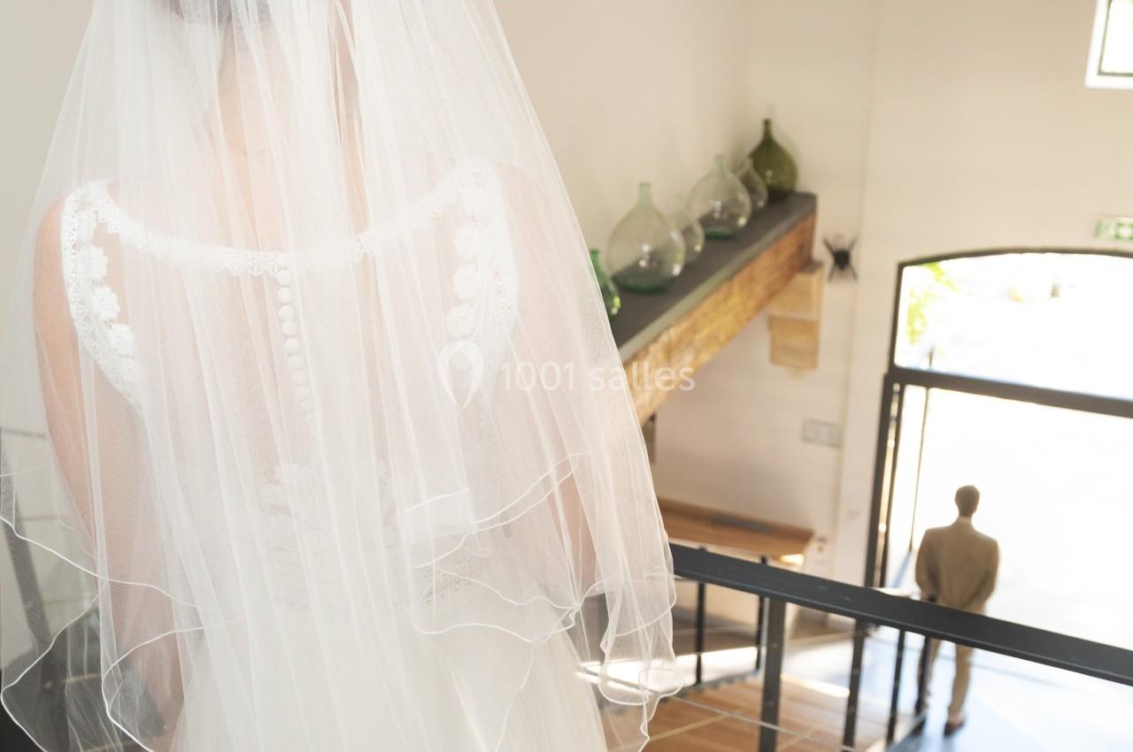 Une mariée en robe blanche et voile descend un escalier en bois, regardant un homme debout en bas.