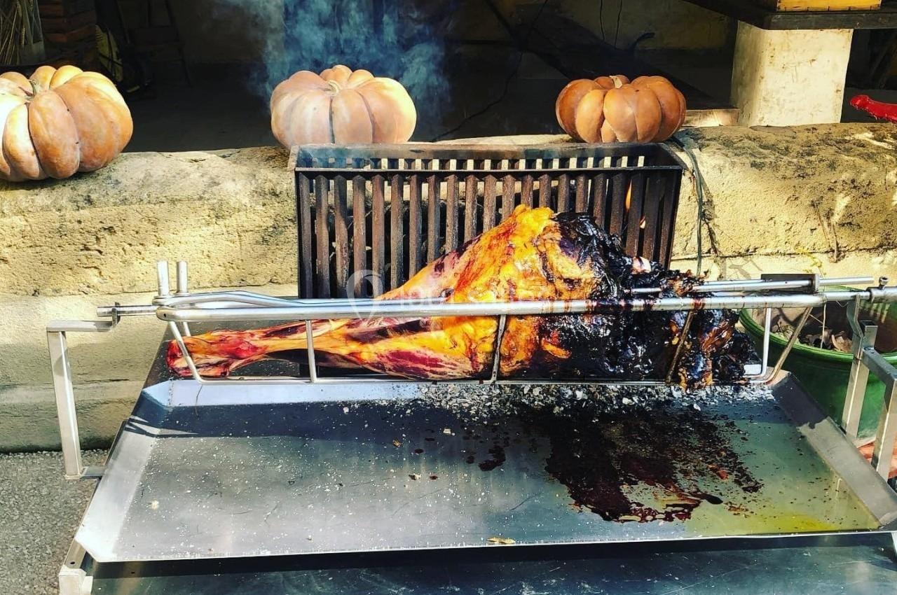 Viande rôtie sur une broche devant un mur en pierre, entourée de citrouilles décoratives.
