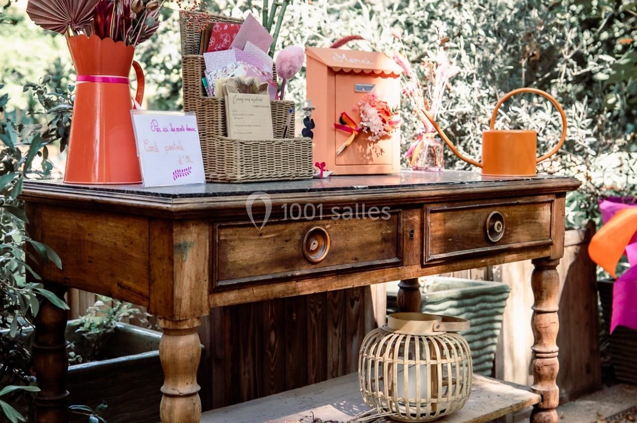 Table en bois avec décorations florales, cartes, arrosoir orange et bouquets séchés, placée dans un jardin ensoleillé.