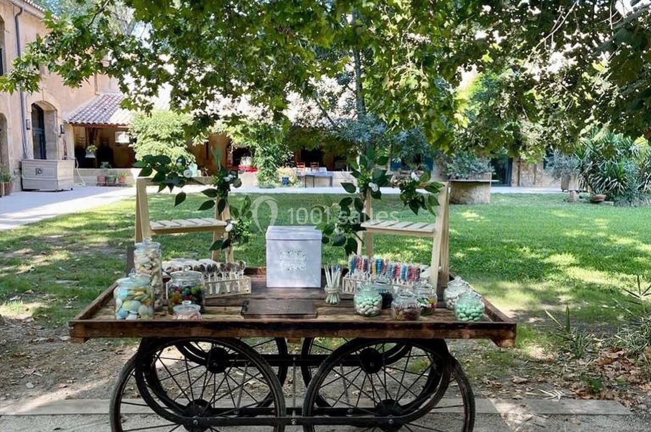 Chariot en bois sur roues, décoré de friandises et de verdure, placé dans un jardin ombragé devant un bâtiment.
