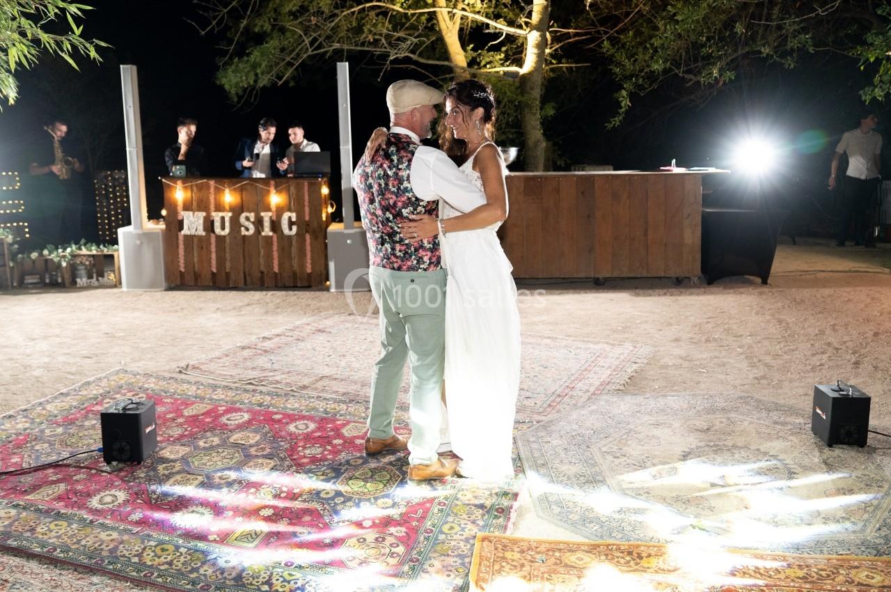 Un couple danse sur des tapis colorés en extérieur, éclairé par des lumières, près d'un bar en bois et d'un panneau ’MUSIC’.