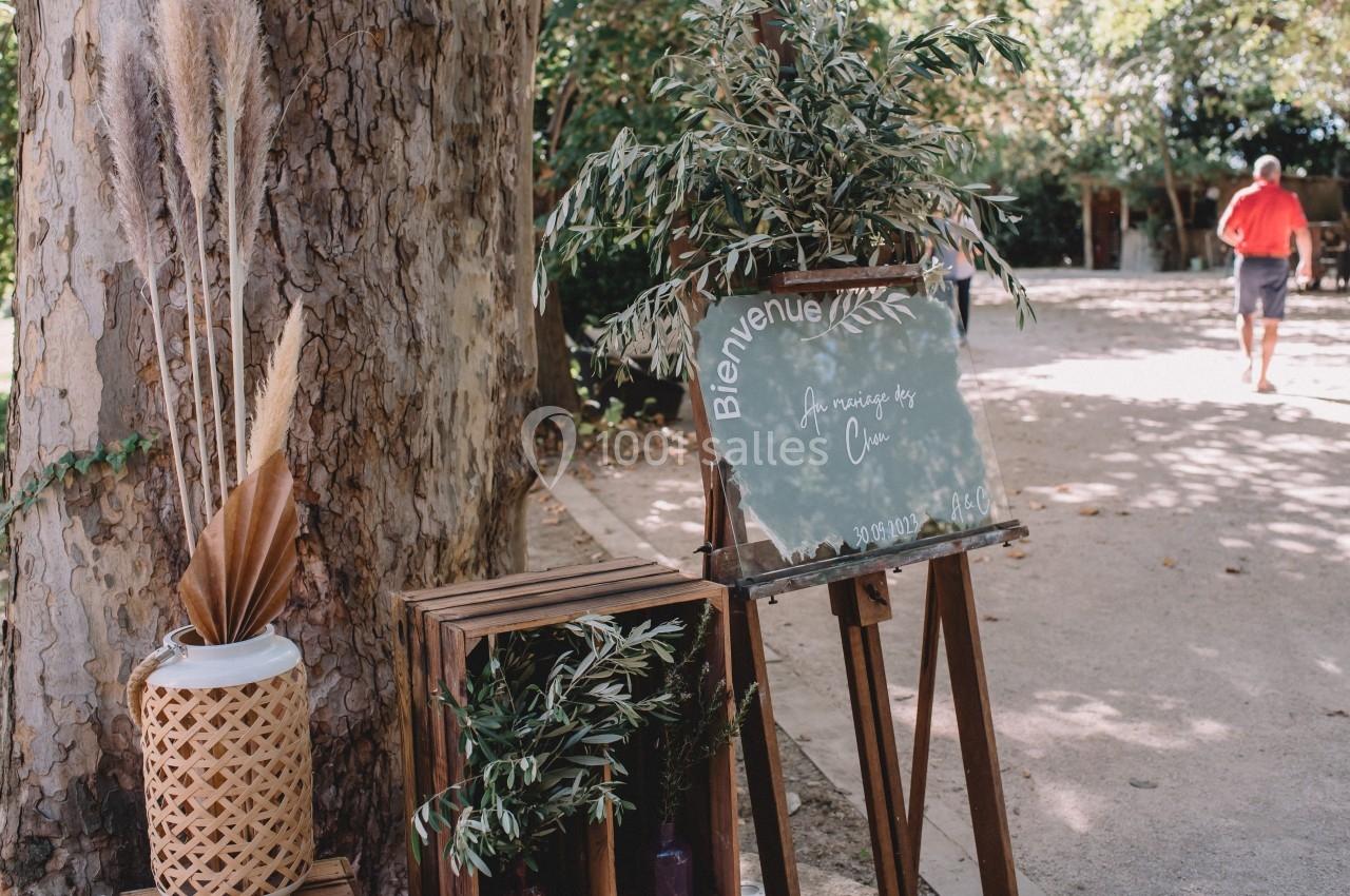 Panneau de bienvenue sur un chevalet entouré de plantes, placé à l'extérieur près d'un arbre.