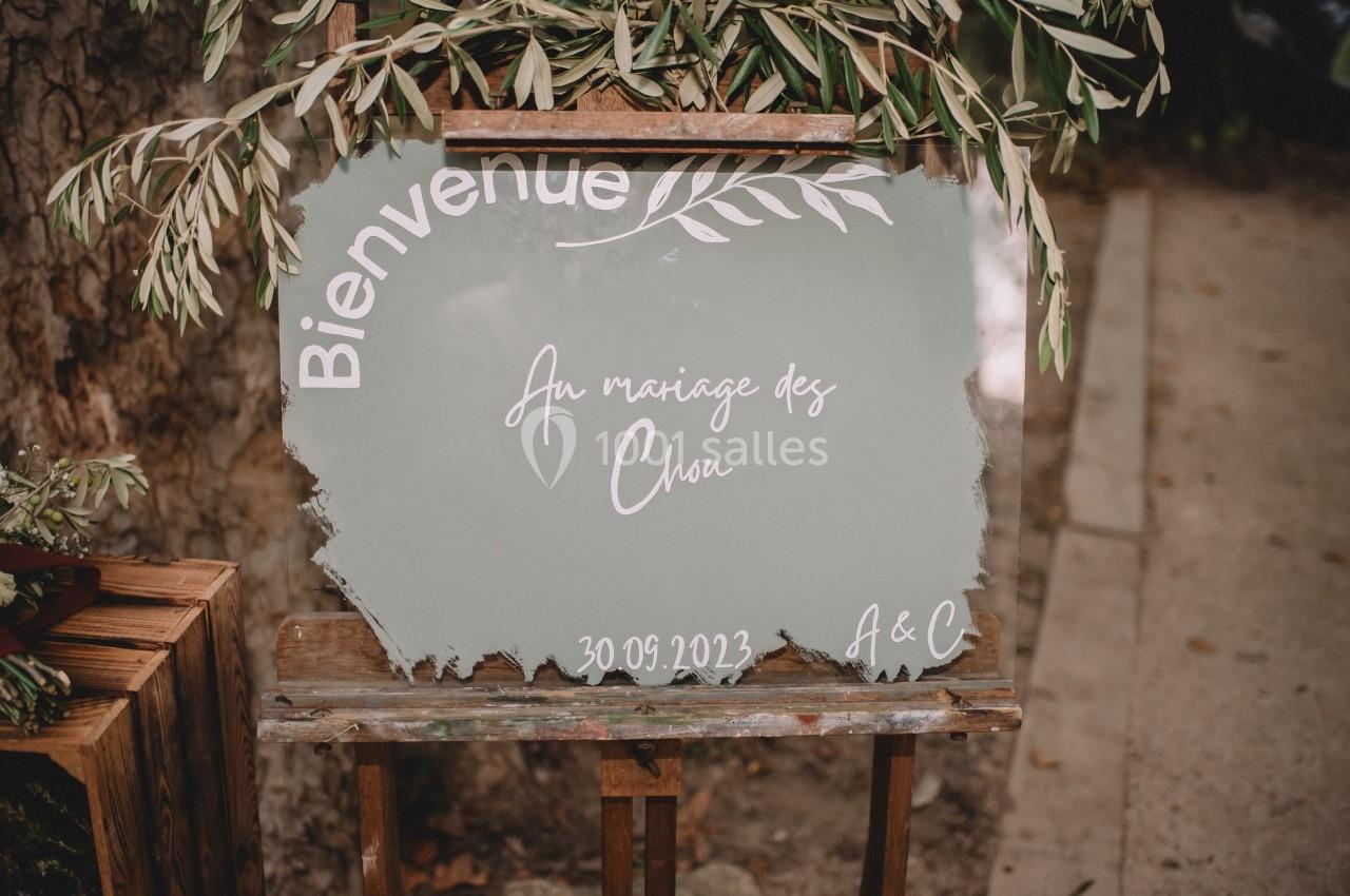 Panneau de bienvenue peint à la main pour un mariage, posé sur un chevalet en bois, entouré de feuillage.