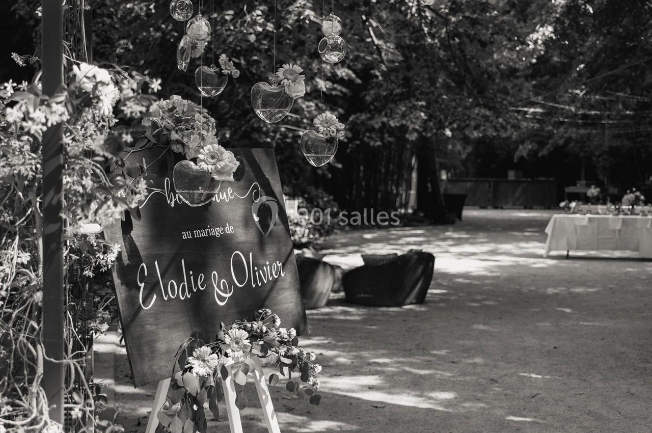 Panneau décoratif annonçant un mariage, entouré de fleurs et suspendu sous des arbres dans un espace extérieur.