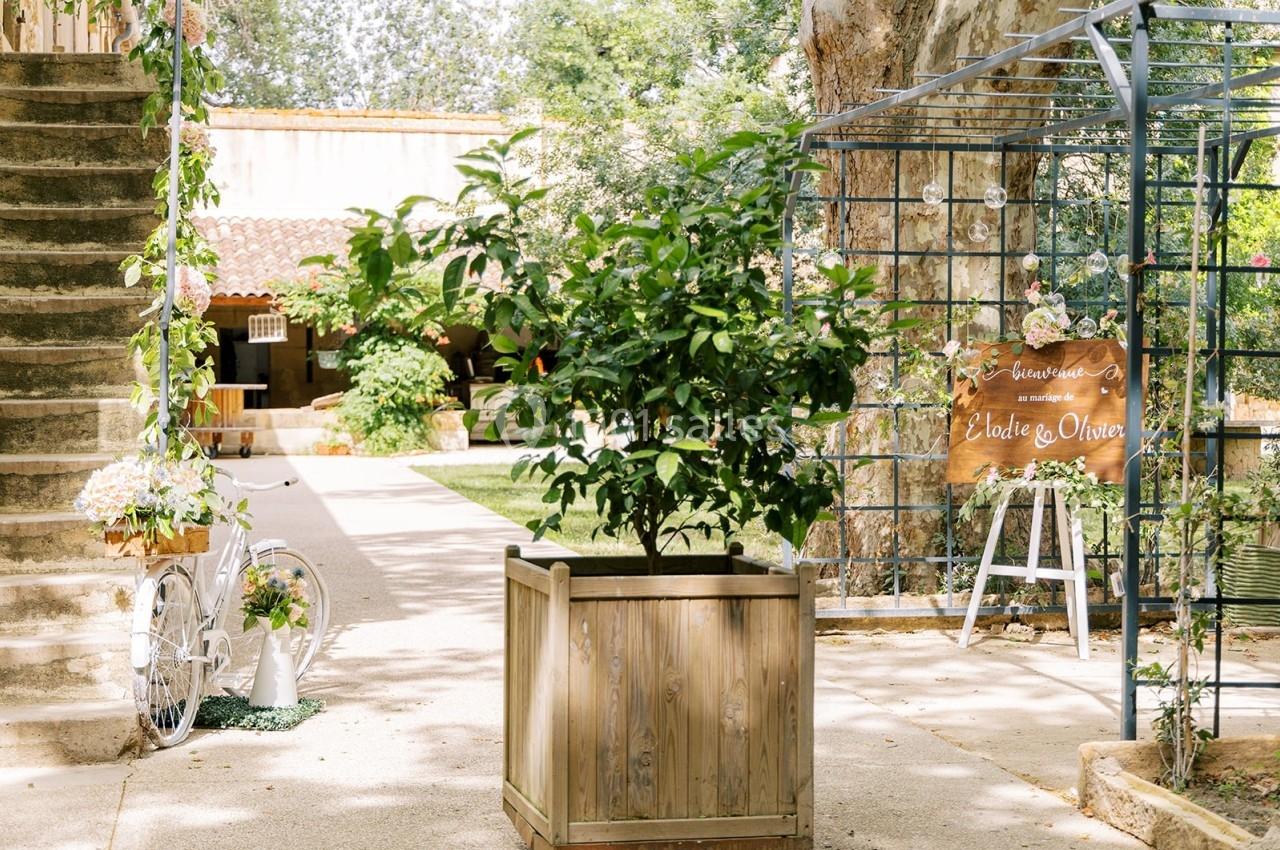Entrée d'un jardin avec un arbre en pot, une arche décorée et une bicyclette blanche près d'un bâtiment en pierre.