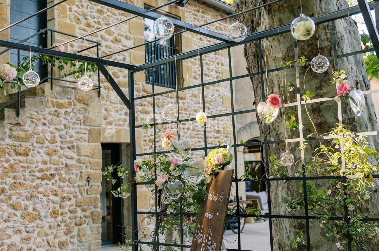 Structure métallique décorée de fleurs et de boules en verre suspendues, devant un mur en pierre et un arbre.