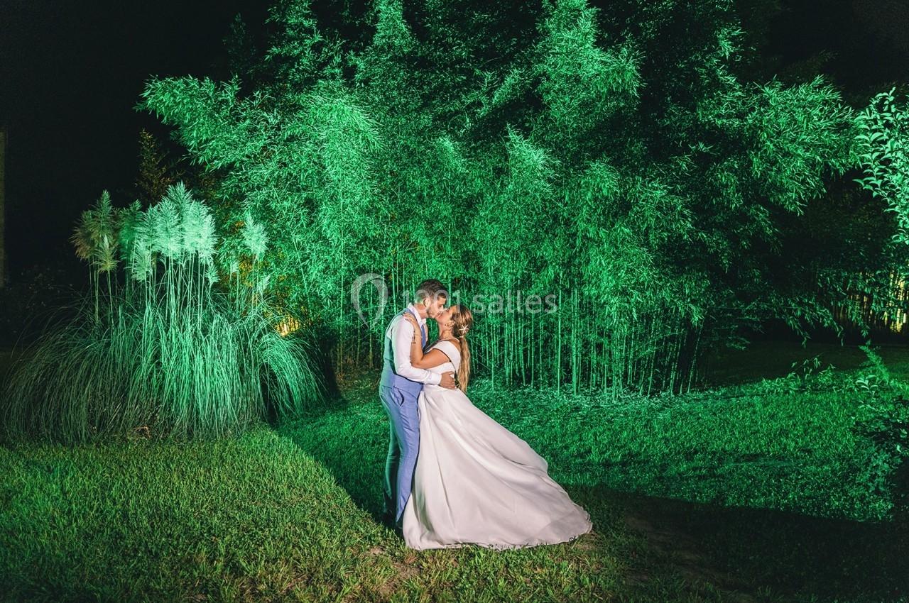 Un couple en tenue de mariage s'embrasse dans un jardin éclairé de lumières vertes, entouré de bambous.