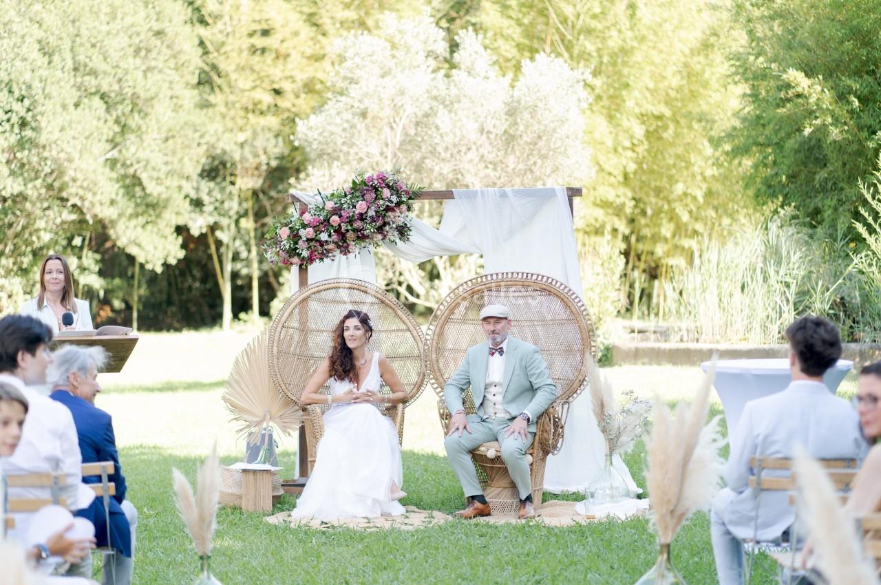 Un couple assis sur des fauteuils en rotin lors d'une cérémonie en plein air, entouré de décorations florales et végétales.