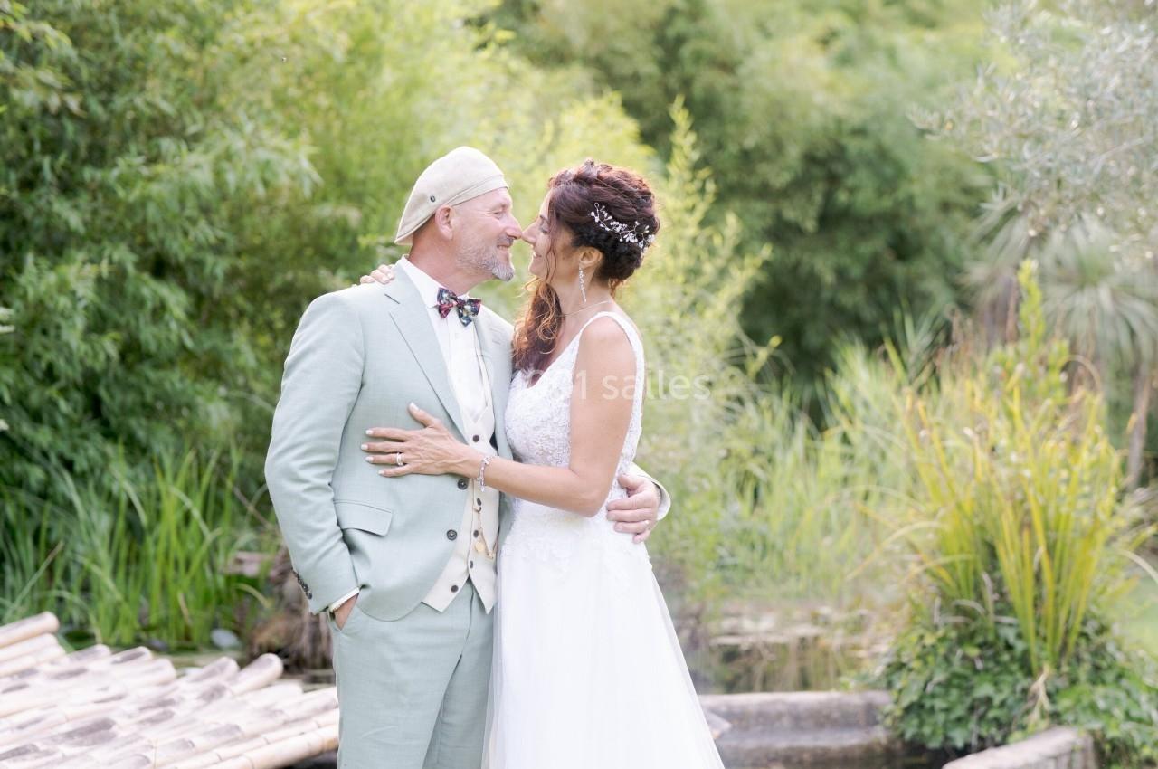Un couple en tenue de mariage se tient debout dans un jardin verdoyant près d'un petit pont en bois.