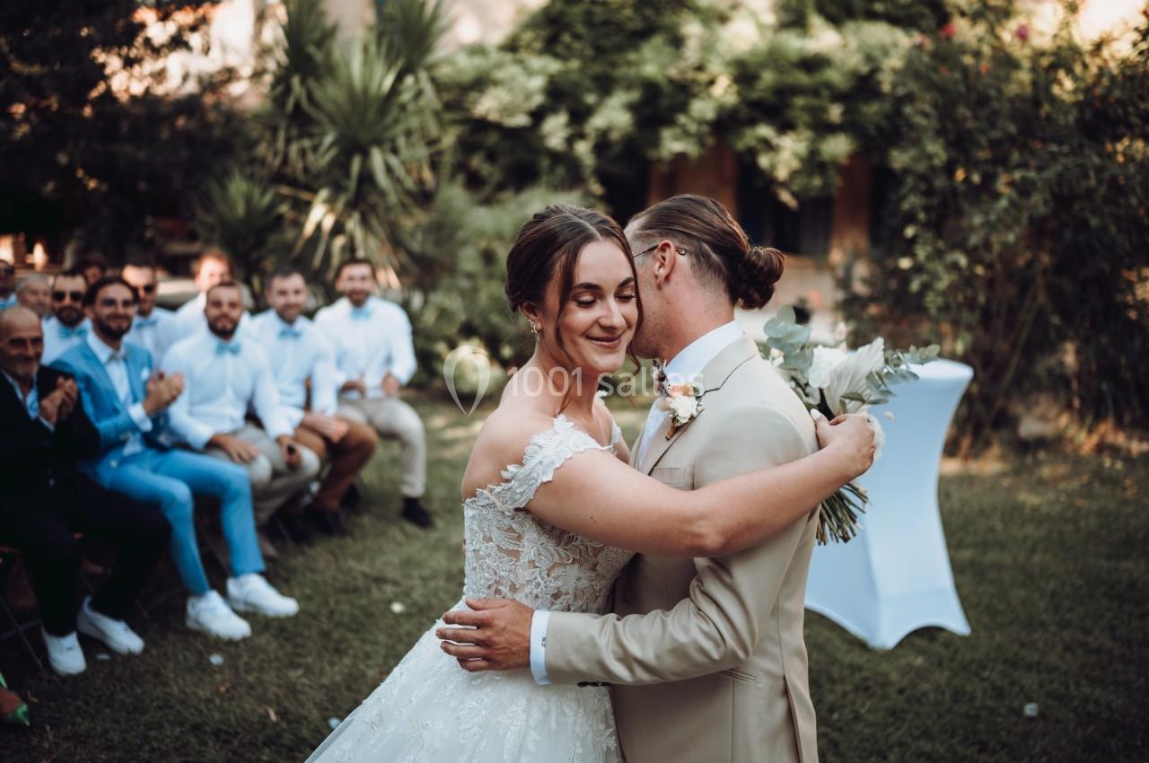 Un couple en tenue de mariage s'enlace dans un jardin, entouré d'invités assis.