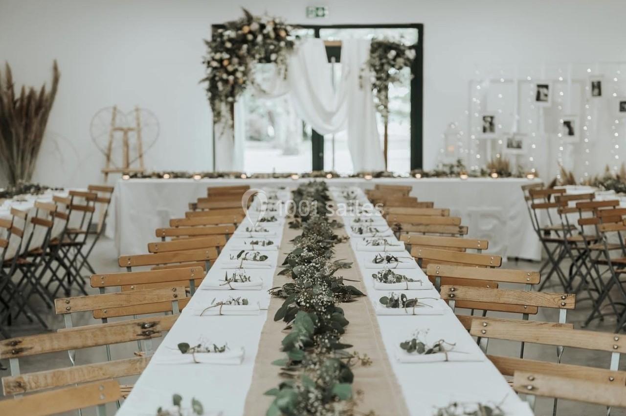 Salle de réception décorée avec des tables alignées, nappes blanches, chemins de table en jute et feuillage, fond floral.
