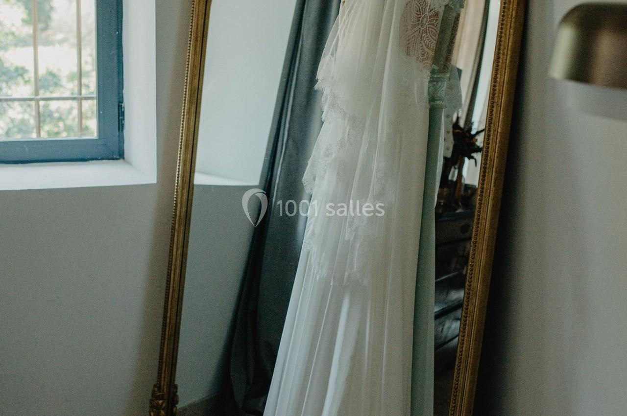 Robe de mariée blanche suspendue à un miroir doré dans une pièce lumineuse avec fenêtre et rideaux.