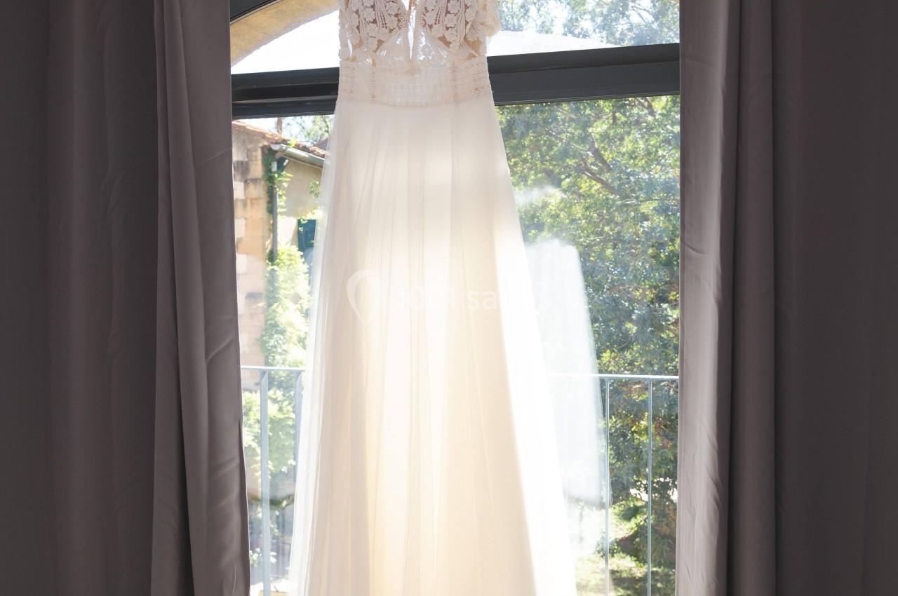 Robe de mariée blanche suspendue à un cintre devant une fenêtre encadrée de rideaux gris.