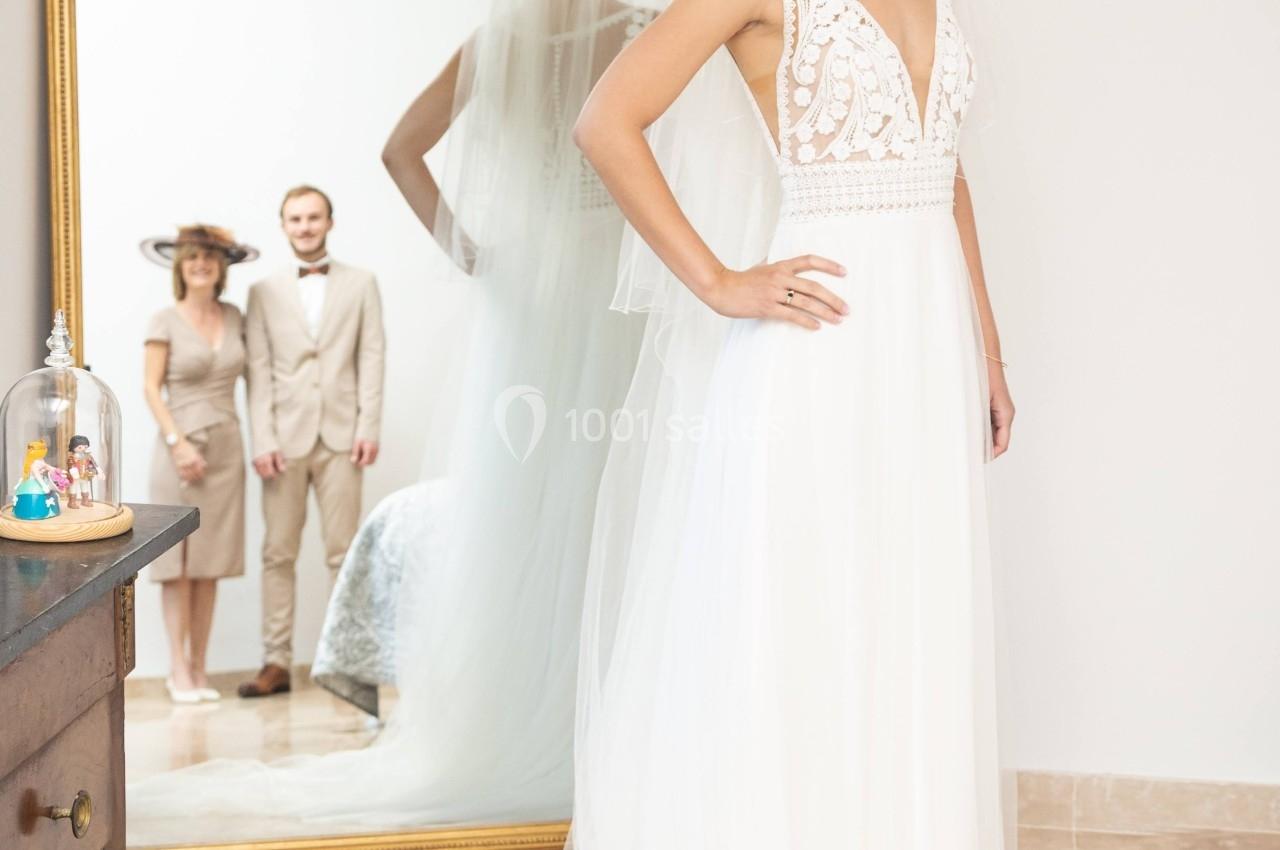 Une mariée en robe blanche se tient devant un miroir, reflétant un couple en arrière-plan dans une pièce lumineuse.