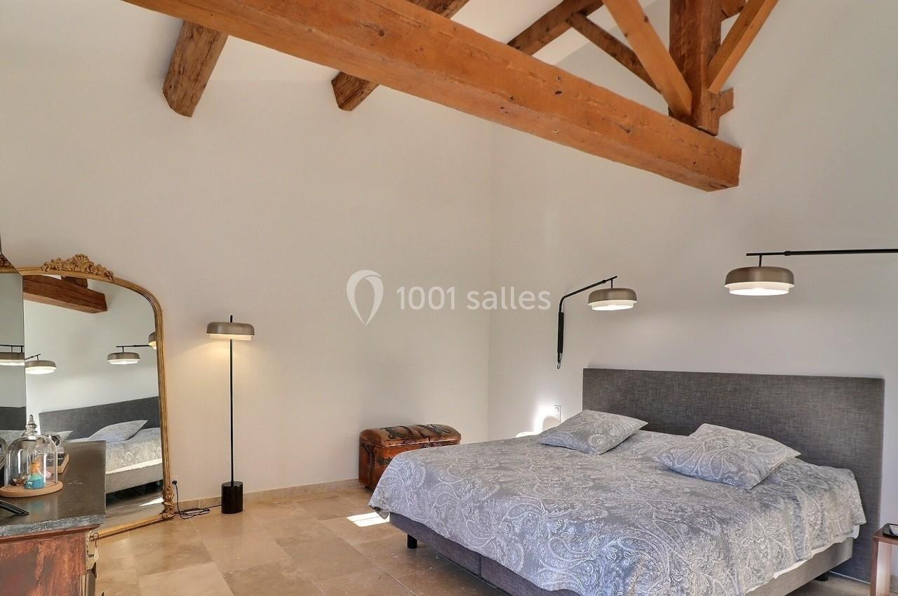Chambre lumineuse avec lit double, poutres apparentes, miroir sur pied, lampes modernes et sol en pierre beige.