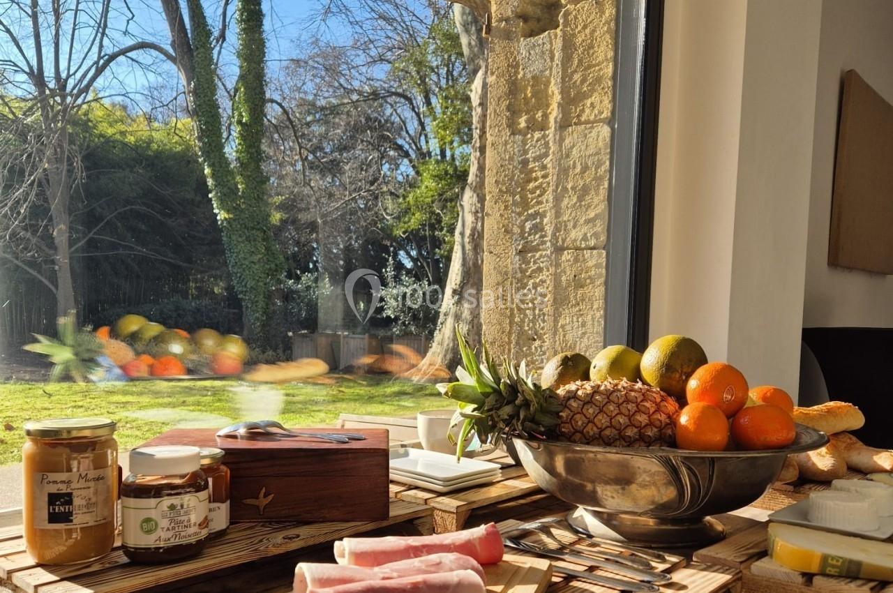 Petit-déjeuner sur une table en bois avec charcuterie, fruits, confitures et vue sur un jardin ensoleillé par une grande…