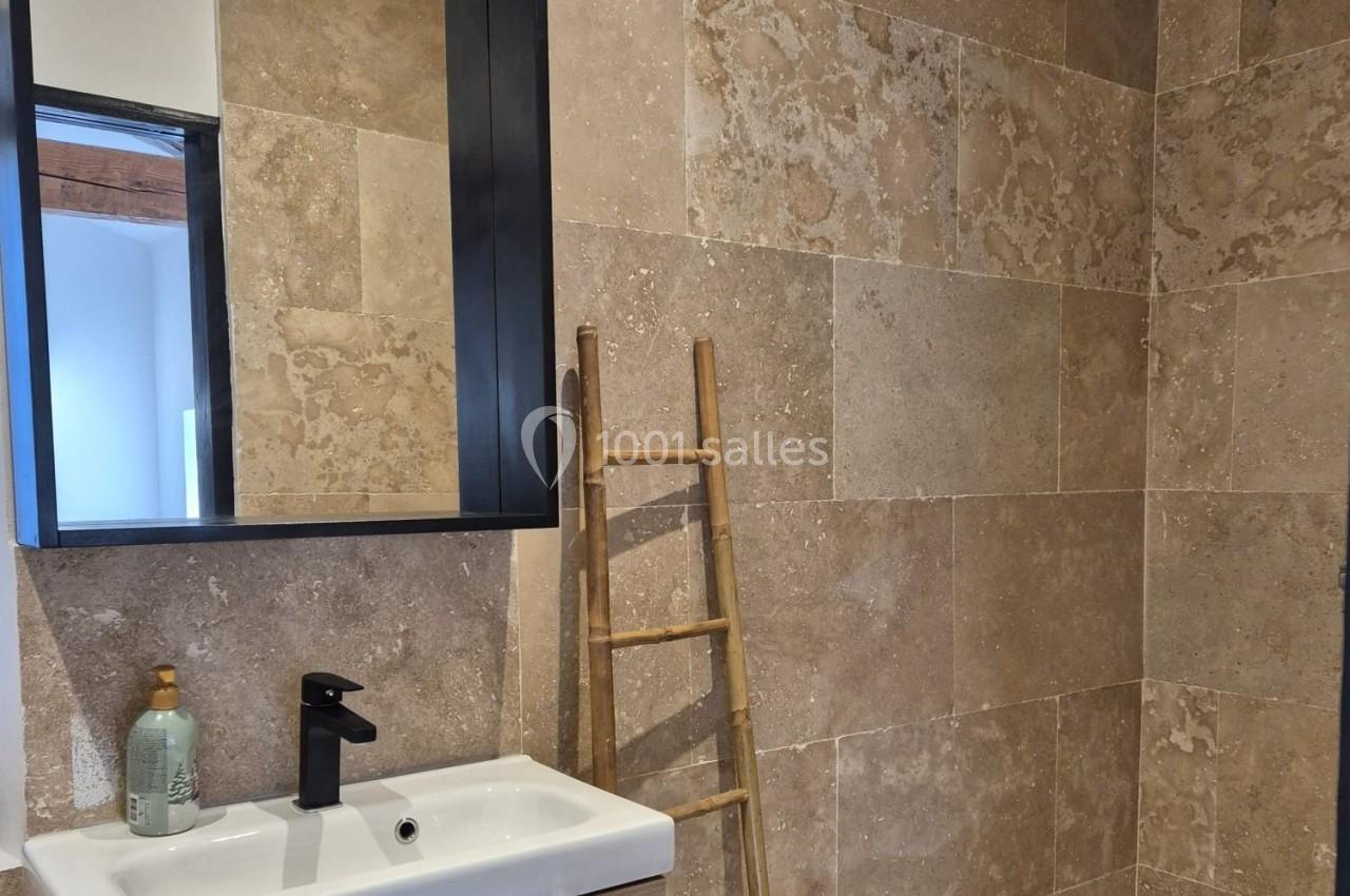 Salle de bain avec lavabo, miroir noir, échelle en bois décorative et murs en carrelage beige texturé.