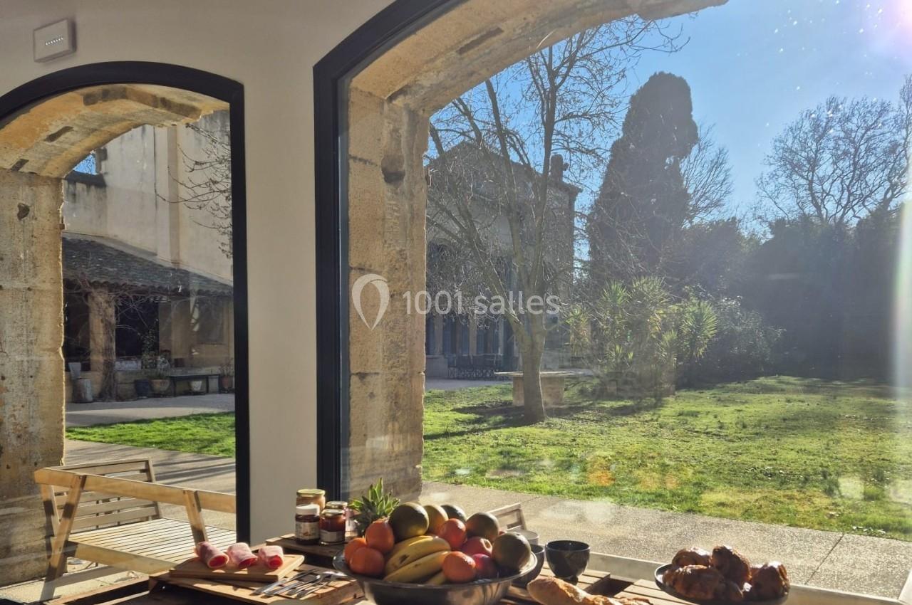 Table en bois garnie de fruits, pain et viennoiseries, près d'une grande baie vitrée donnant sur un jardin ensoleillé.
