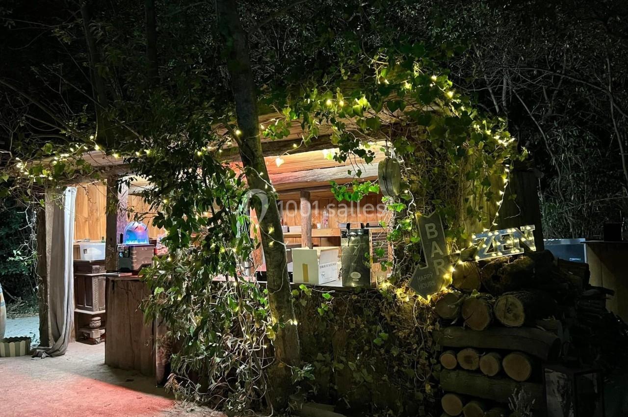 Un bar extérieur en bois décoré de guirlandes lumineuses et entouré de végétation, éclairé de nuit.