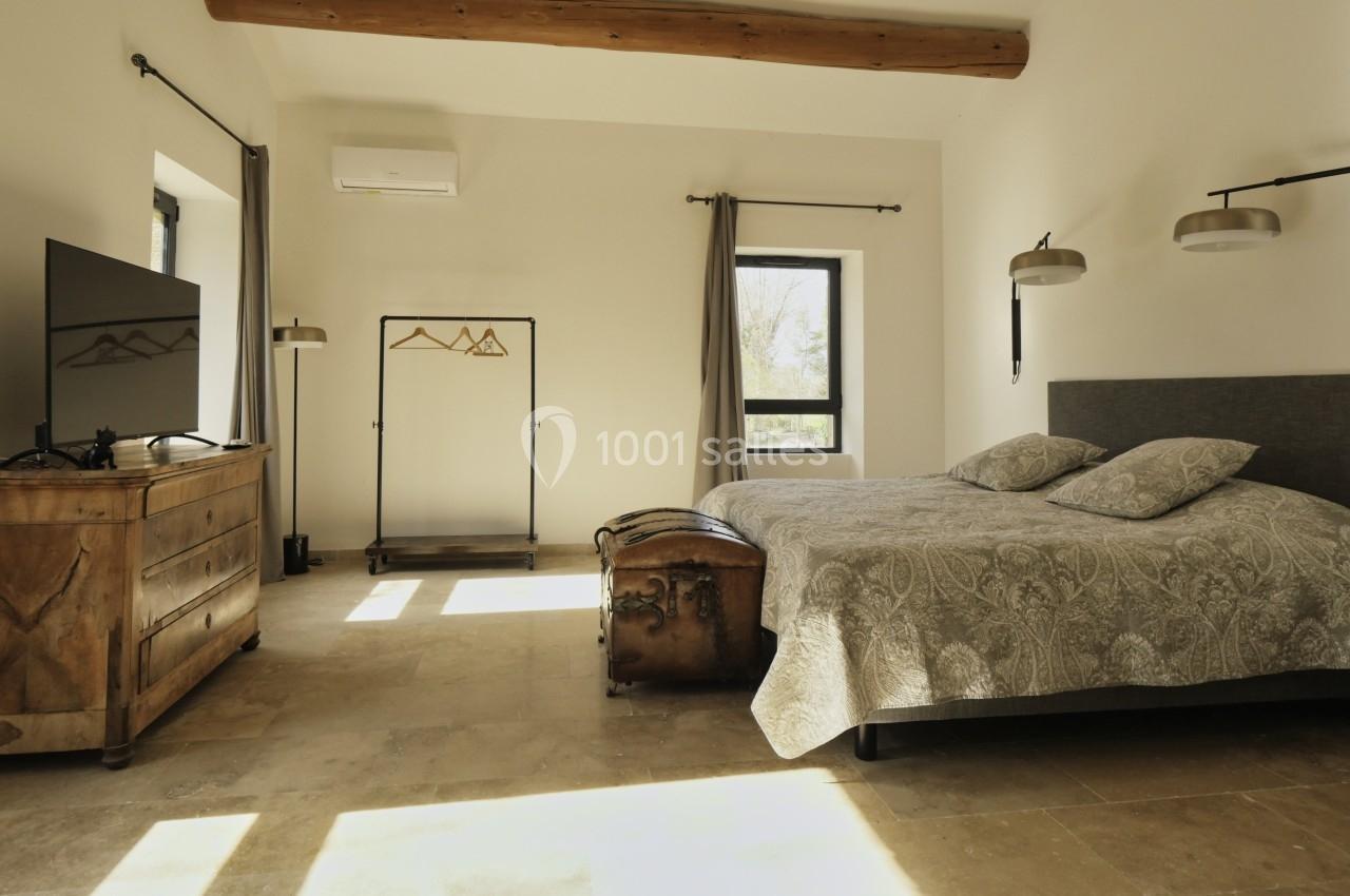 Chambre lumineuse avec lit double, commode en bois, portant à vêtements, télévision et poutre apparente au plafond.