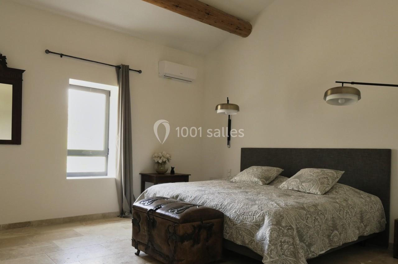 Chambre lumineuse avec un lit double, des lampes suspendues, un coffre en bois et une fenêtre avec rideaux gris.