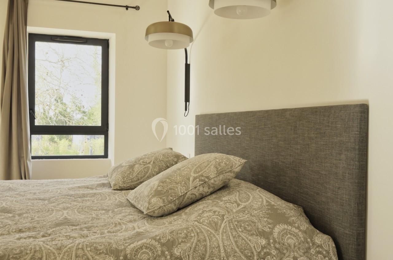 Lit double avec tête de lit grise, draps à motifs, deux lampes murales et une fenêtre avec rideaux beiges.