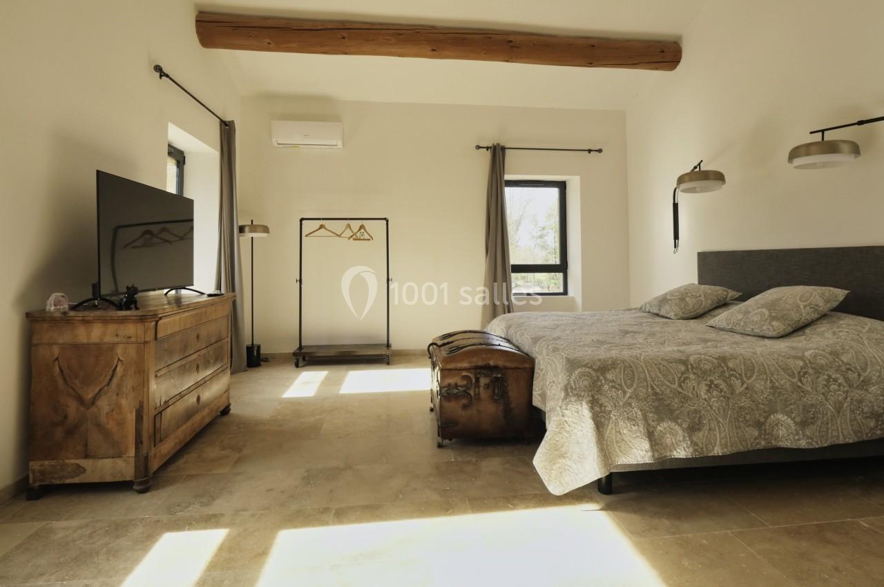 Chambre lumineuse avec lit double, commode en bois, portant à vêtements, télévision et poutre apparente au plafond.