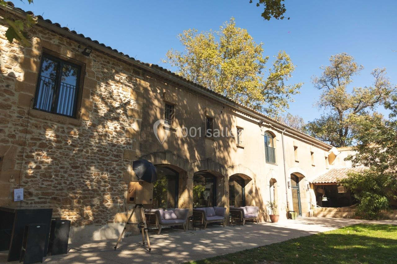 Façade en pierre d'un bâtiment ancien avec terrasse aménagée, entouré d'arbres sous un ciel dégagé.