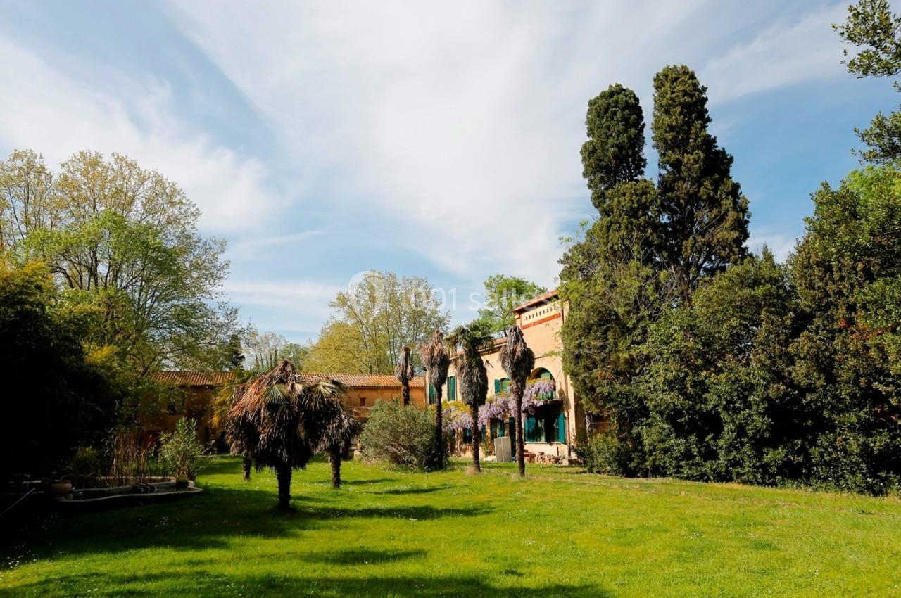 Jardin verdoyant avec pelouse, arbres, palmiers et bâtiment en arrière-plan, sous un ciel partiellement nuageux.