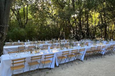 Tables rondes dressées avec nappes blanches et chaises en bois, disposées en extérieur sous des arbres verdoyants.