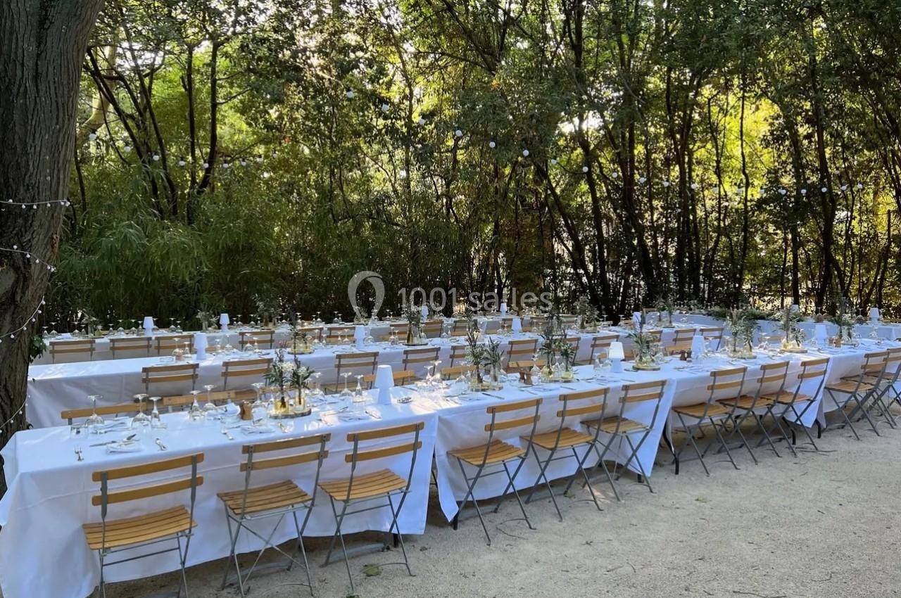 Tables longues dressées avec nappes blanches et chaises en bois, installées en extérieur sous des arbres.