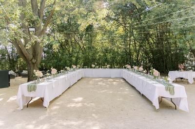 Tables rondes dressées avec nappes blanches et chaises en bois, disposées en extérieur sous des arbres verdoyants.