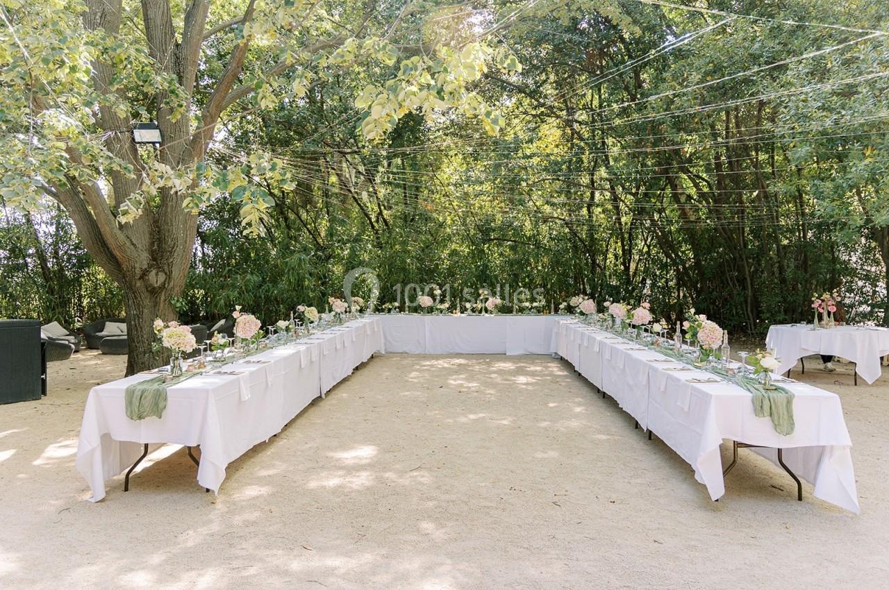 Tables disposées en U avec nappes blanches et décorations florales, installées en extérieur sous des arbres.