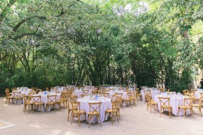 Tables rondes dressées avec nappes blanches et chaises en bois, disposées en extérieur sous des arbres verdoyants.