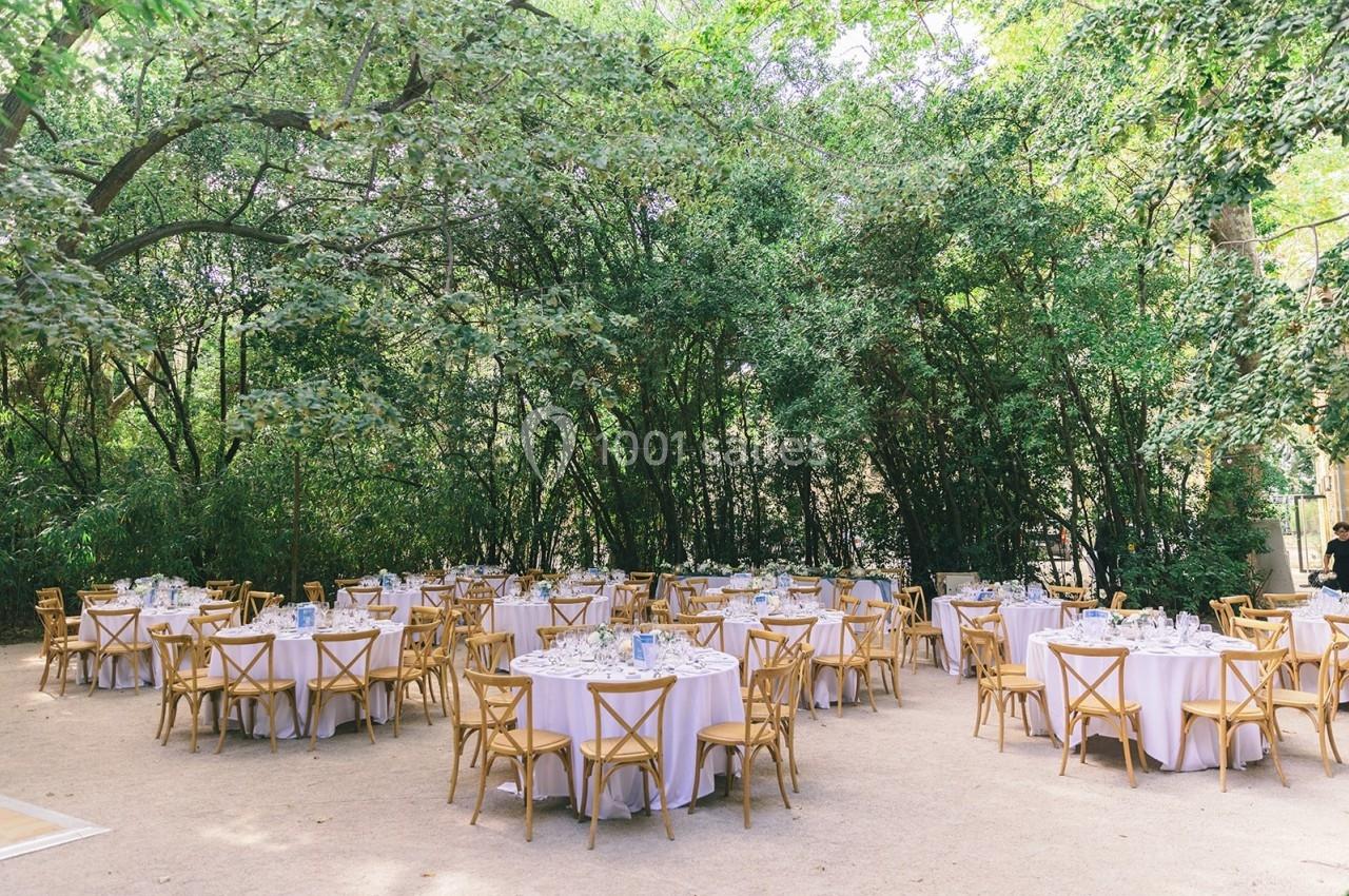 Tables rondes dressées avec nappes blanches et chaises en bois, disposées en extérieur sous des arbres verdoyants.