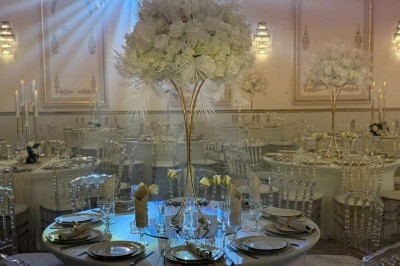 Table ronde élégamment dressée avec nappes blanches, chaises transparentes, centre de table floral et bougies hautes.