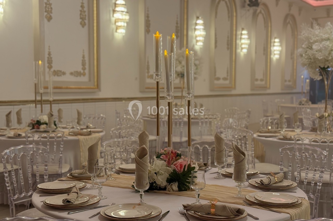 Salle de réception élégante avec tables rondes dressées, chandeliers et décorations florales.