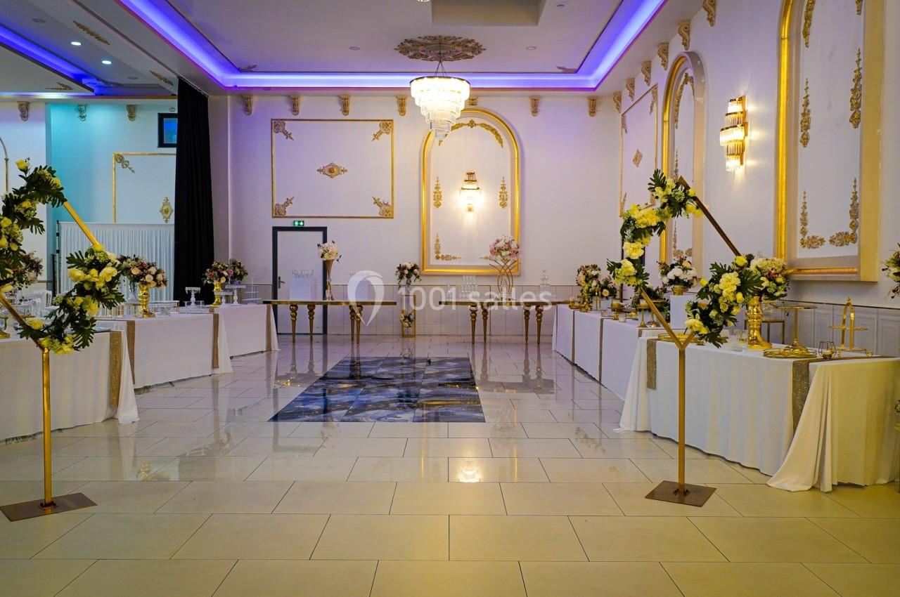 Salle de réception élégante avec décorations florales, tables dressées et éclairage violet au plafond.