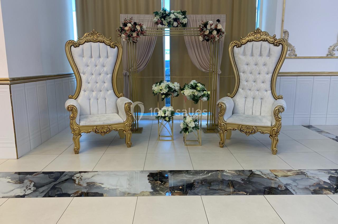 Deux fauteuils dorés ornés de motifs, entourés de fleurs blanches et roses, devant un rideau beige.