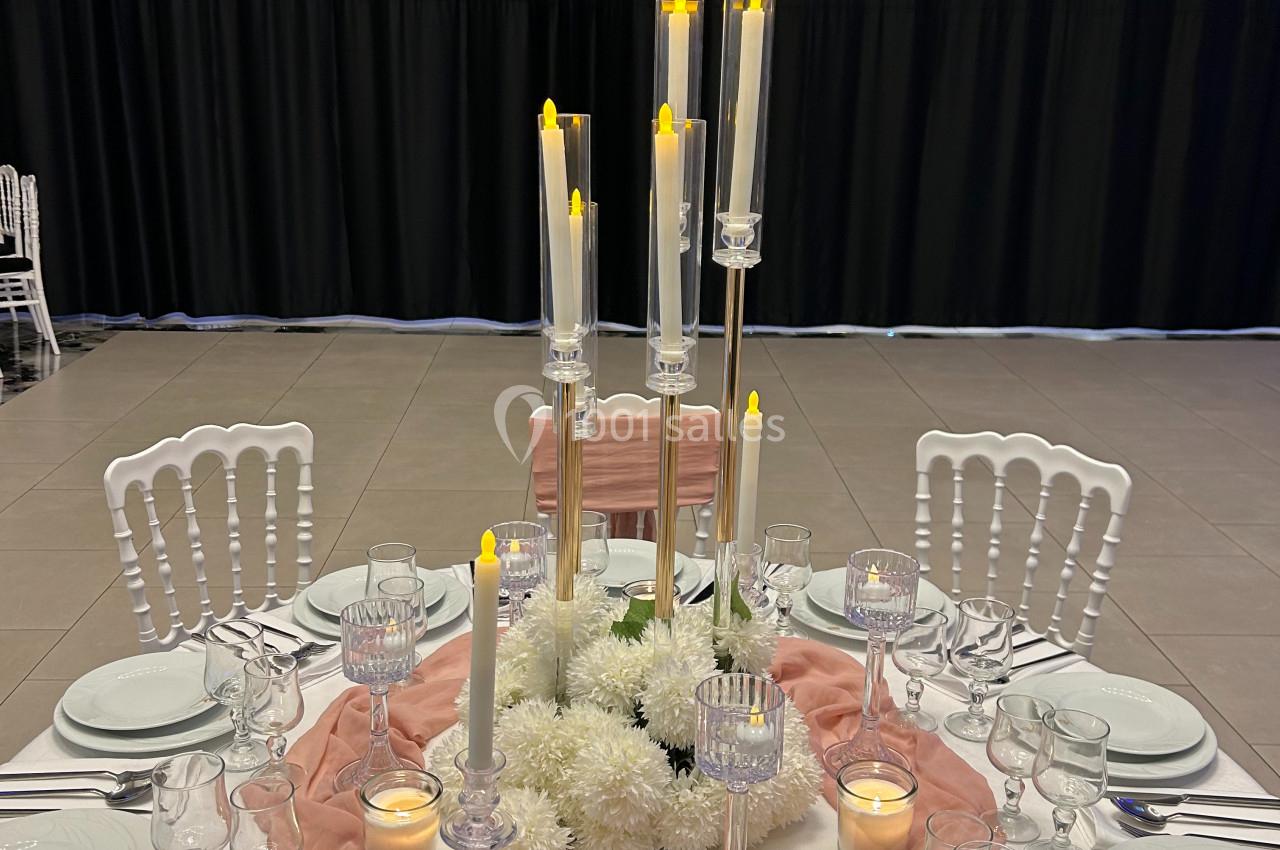Table élégamment dressée avec nappes blanches, chemin de table rose, chandeliers hauts et décorations florales blanches.