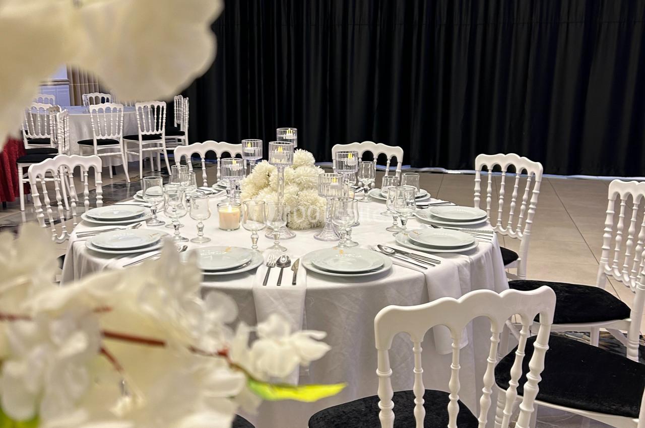 Table dressée avec nappes blanches, vaisselle élégante et centre de table floral, entourée de chaises blanches.