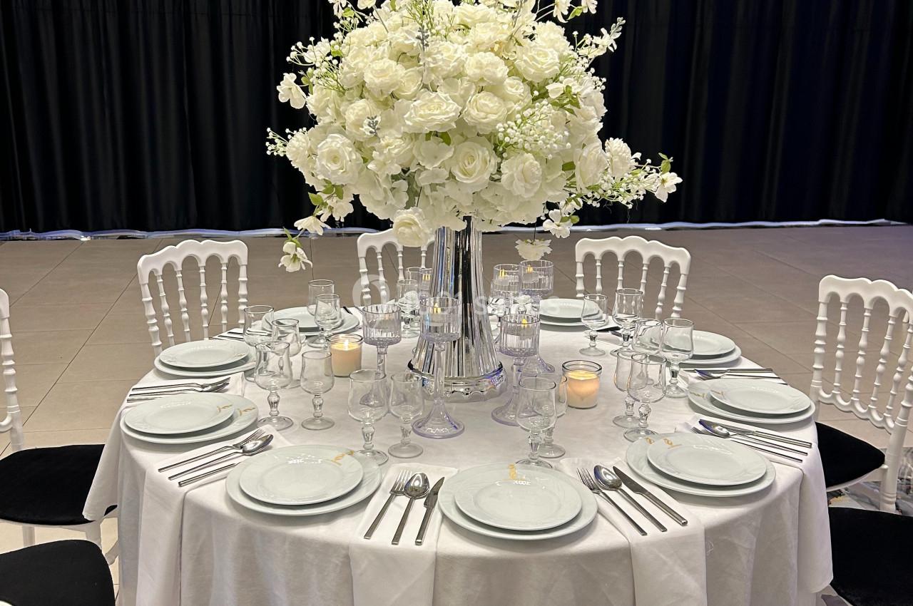 Table ronde élégamment dressée avec une nappe blanche, des couverts alignés et un grand bouquet de fleurs blanches au centre.