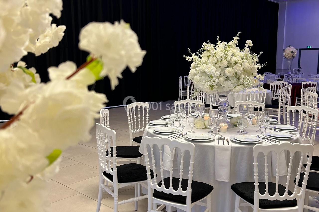Salle de réception élégante avec tables rondes dressées, décorées de fleurs blanches et chaises blanches.