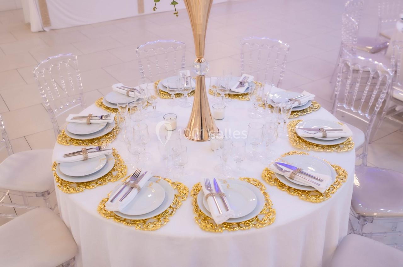 Table ronde décorée pour un événement, avec nappes blanches, assiettes dorées, couverts et centre de table floral.