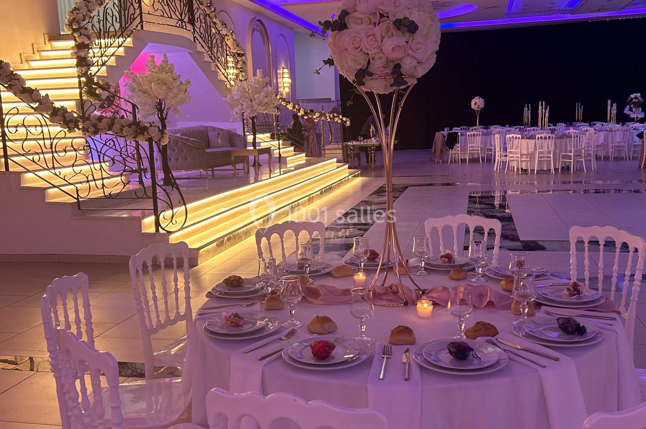 Salle de réception élégante avec table dressée, décor floral et escalier illuminé en arrière-plan.