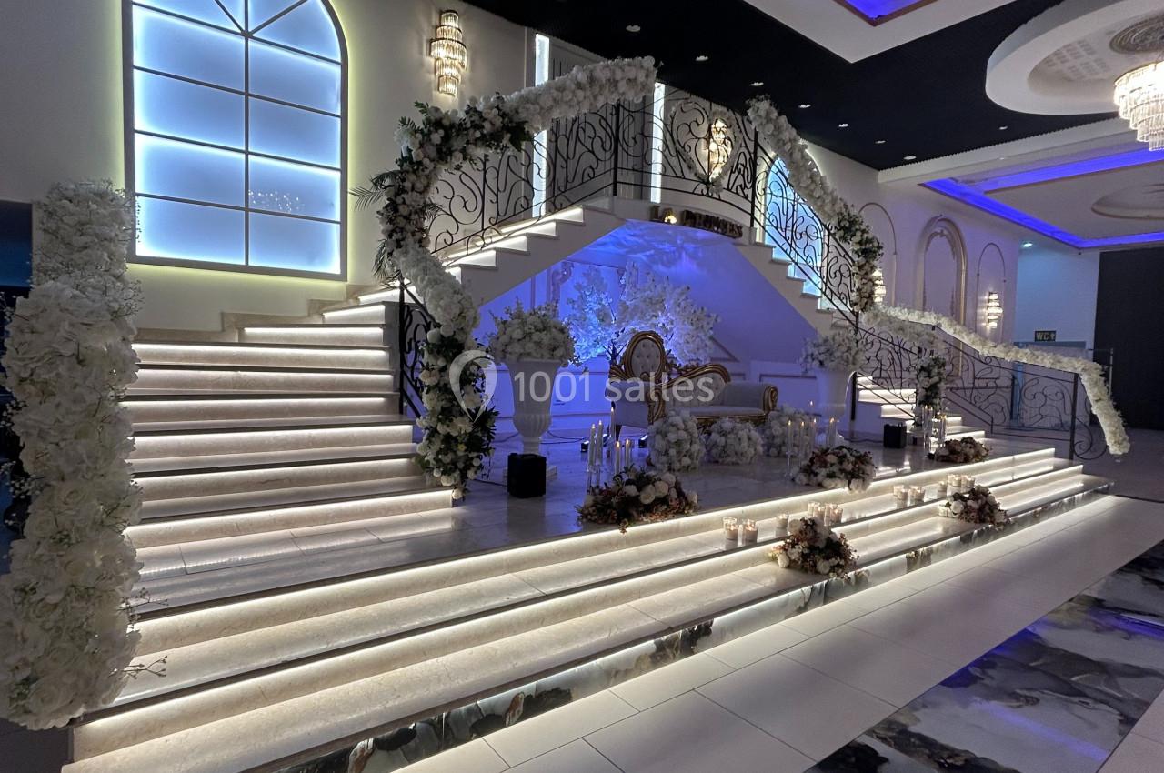 Escalier décoré de fleurs blanches illuminé par des LED, menant à une scène ornée dans une salle de réception élégante.