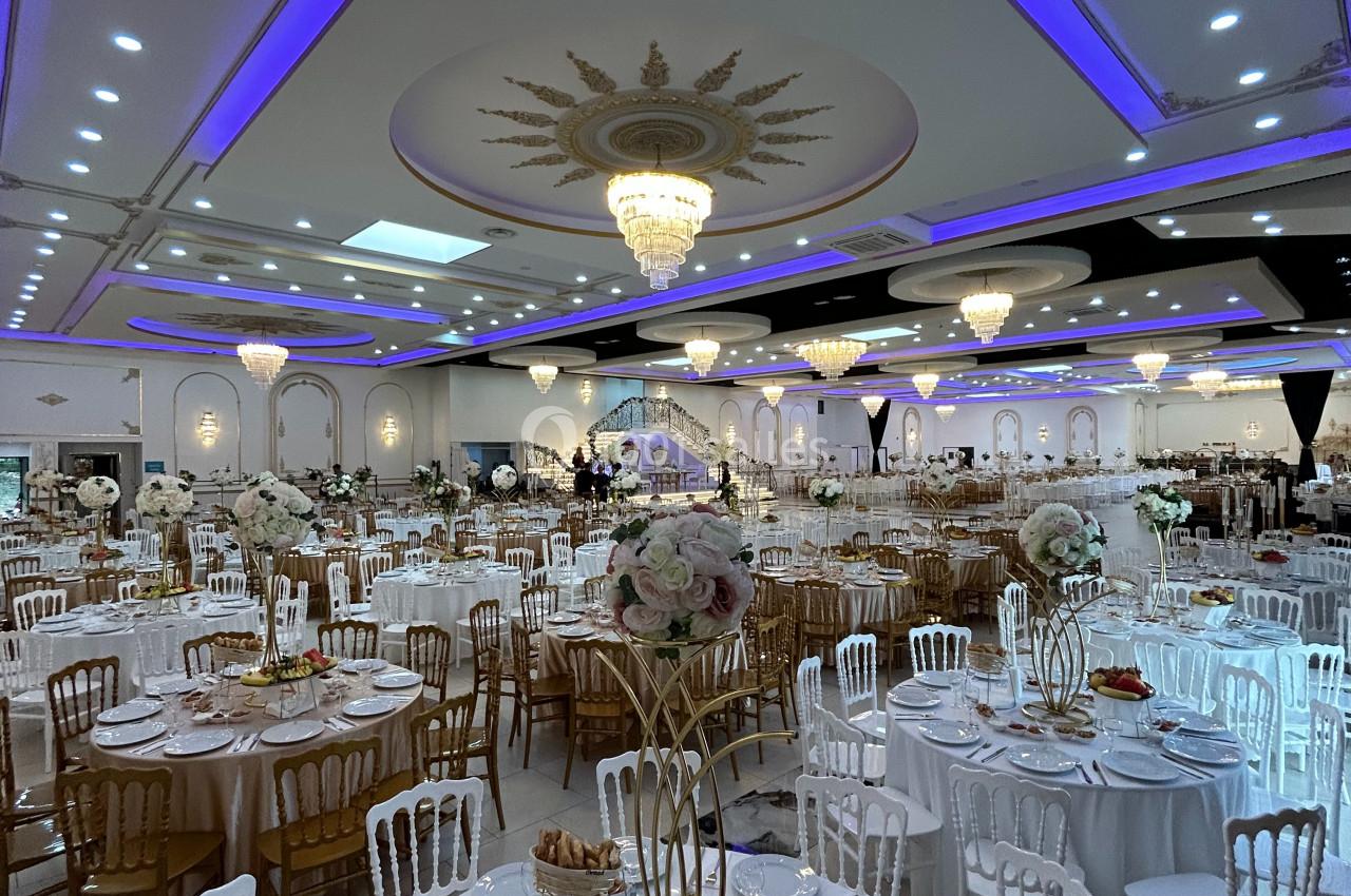 Salle de réception élégante avec tables dressées, chaises blanches et dorées, éclairage violet et lustres imposants.